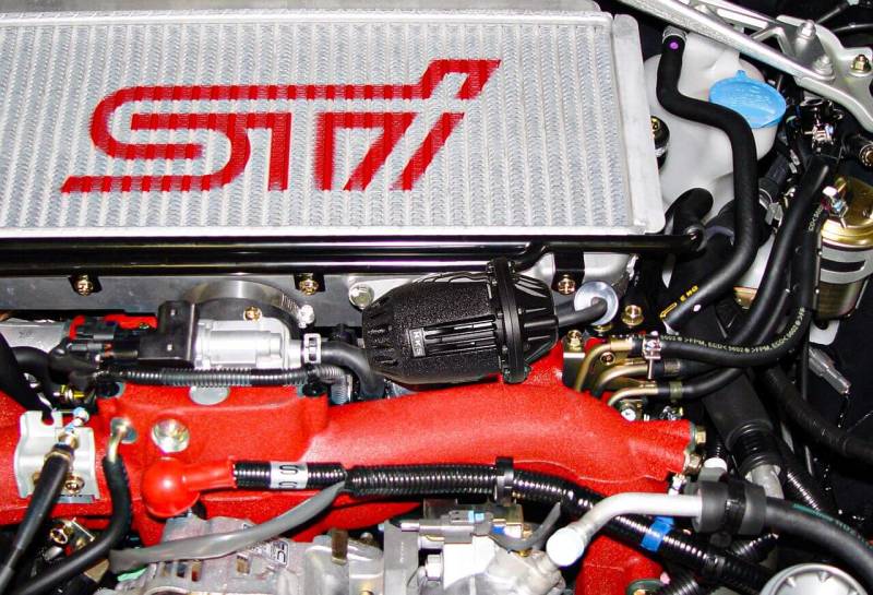 A close-up image of an HKS Super SQV4 Black Edition GDB for EJ205/EJ255/EJ257 engines, showing the product's placement in an automotive setting, specifically focusing on the product's design and the surrounding engine components.